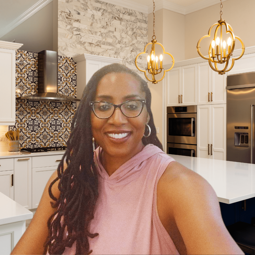 A smiling woman with long, dark dreadlocks and glasses poses in a modern kitchen with white cabinetry, gold pendant lighting, and a bold patterned backsplash behind the stove. She wears a sleeveless pink hoodie and has a warm, friendly expression. The kitchen features a sleek design with stainless steel appliances, a marble accent wall, and a large island with a glossy white countertop.