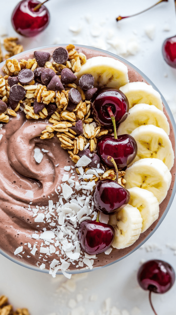 A creamy chocolate-covered cherry smoothie bowl topped with fresh cherries, banana slices, granola, chocolate chips, and shredded coconut. The thick, swirled smoothie base provides a rich and indulgent texture, while the toppings add a mix of crunch and sweetness. The vibrant colors and fresh ingredients create a visually appealing and nourishing breakfast or dessert option.