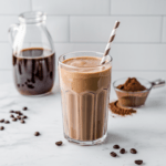 A creamy mocha coffee protein smoothie served in a tall glass with a striped brown and white straw. The smoothie has a rich, frothy texture and a deep coffee-chocolate color. In the background, a glass pitcher of brewed coffee, a scoop of cocoa powder, and scattered coffee beans on a marble countertop highlight the drink's key ingredients.