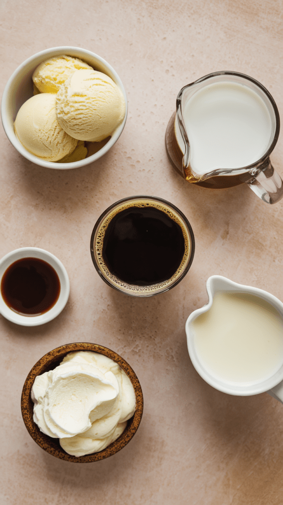 A flat lay of ingredients for a mudslide mocktail, arranged on a neutral-toned surface. The spread includes a bowl of vanilla ice cream, a glass of brewed coffee, a small dish of vanilla extract, a bowl of whipped cream, and two pitchers—one with milk and the other with a creamy liquid. The ingredients are neatly arranged, showcasing the rich and indulgent components of this dessert-inspired drink.