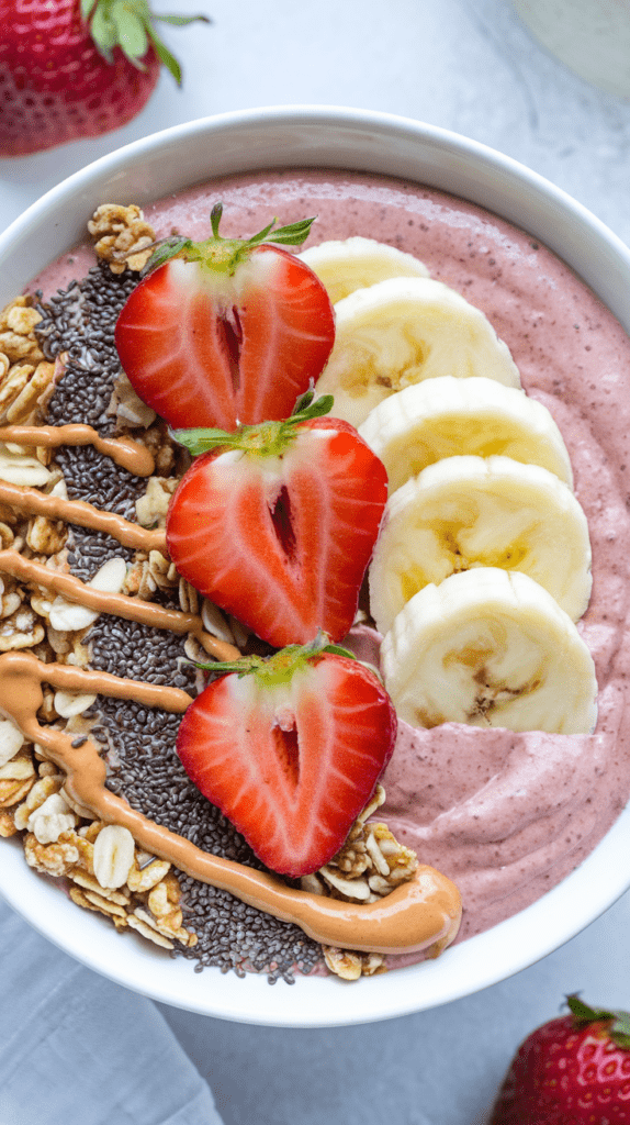 A creamy strawberry banana protein smoothie bowl topped with sliced fresh strawberries, banana slices, granola, chia seeds, and a drizzle of peanut butter. The smooth, thick texture of the smoothie contrasts with the crunchy toppings, creating a balanced and nutritious meal. The bright colors and fresh ingredients make this bowl both visually appealing and packed with flavor.