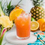 A vibrant tropical punch mocktail served in a textured glass filled with ice, garnished with a fresh orange slice. The drink has a bright orange-pink hue, with bubbles visible on the surface. In the background, fresh tropical fruits like pineapple, oranges, lemons, and limes add to the summery aesthetic, while a colorful floral napkin rests nearby.